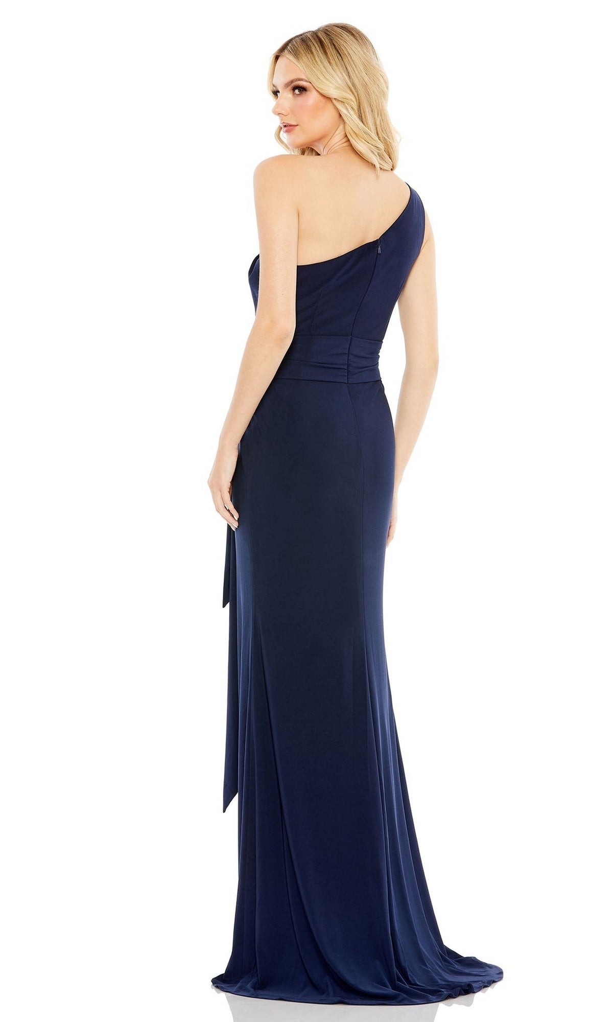 A woman models the Mac Duggal 26581 Belted Long Jersey Formal Dress in navy blue, standing slightly turned from the camera against a white background to elegantly display its one-shoulder, floor-length design.